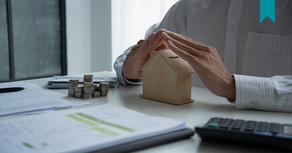Landlord and Tenant insurance being represented by hands covering wood block shaped home on desk with documents surrounding.